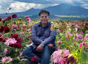 Alejandro Lira sentado entre flores, con montañas al fondo.