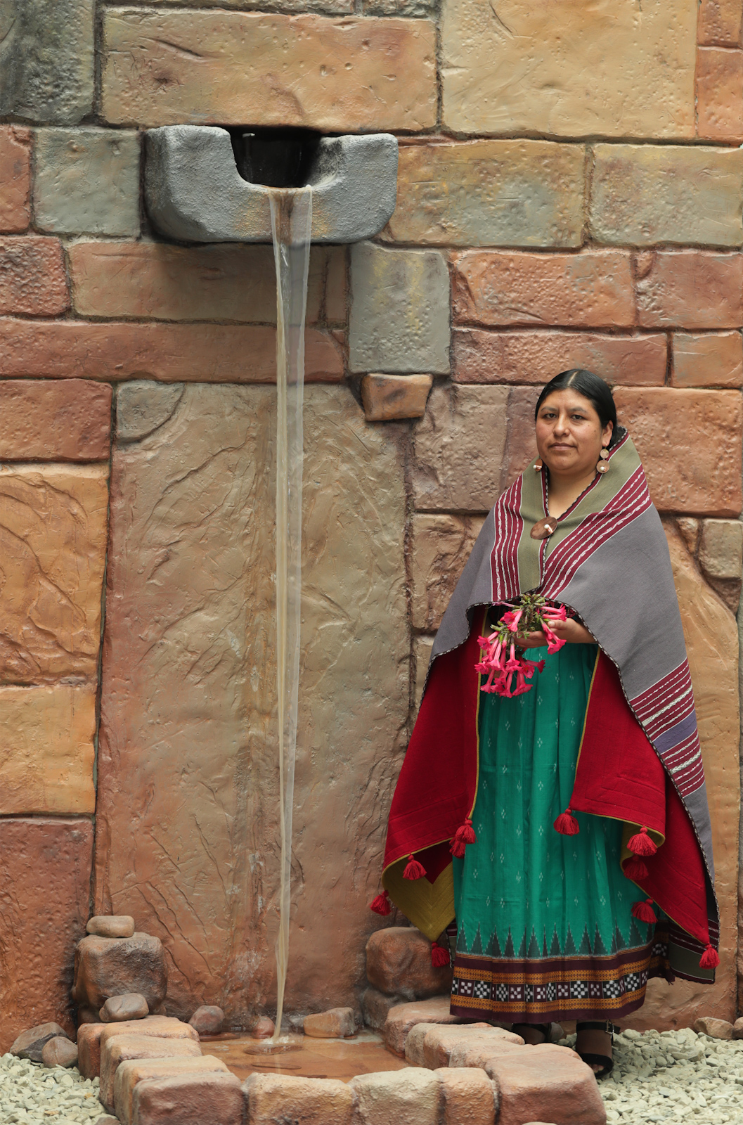 Elvira Espejo Ayca de pie junto a un muro de piedra, con prendas textiles y una falda verde.