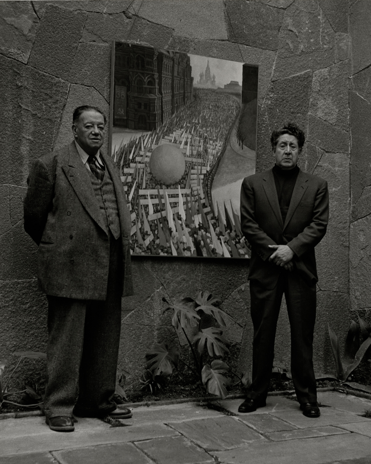 Diego Rivera y David Alfaro Siqueiros de pie junto a una pintura, en una fotografía en blanco y negro de Rodrigo Moya.